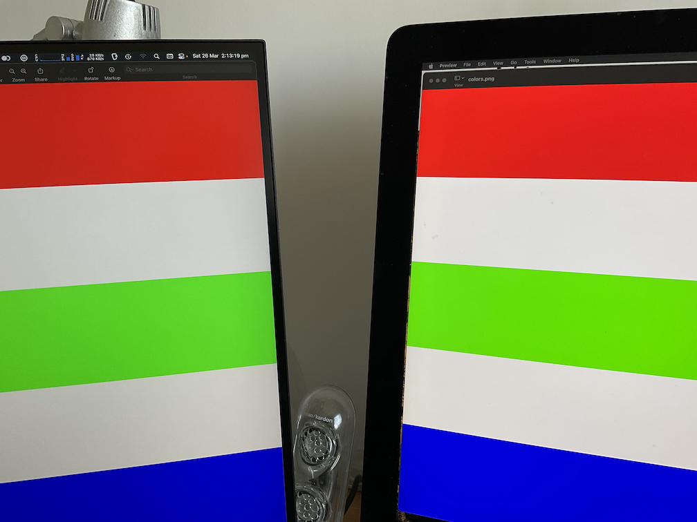 Photo of M28U and iMac next to each other, both showing an image with stripes of white, blue, green, and red